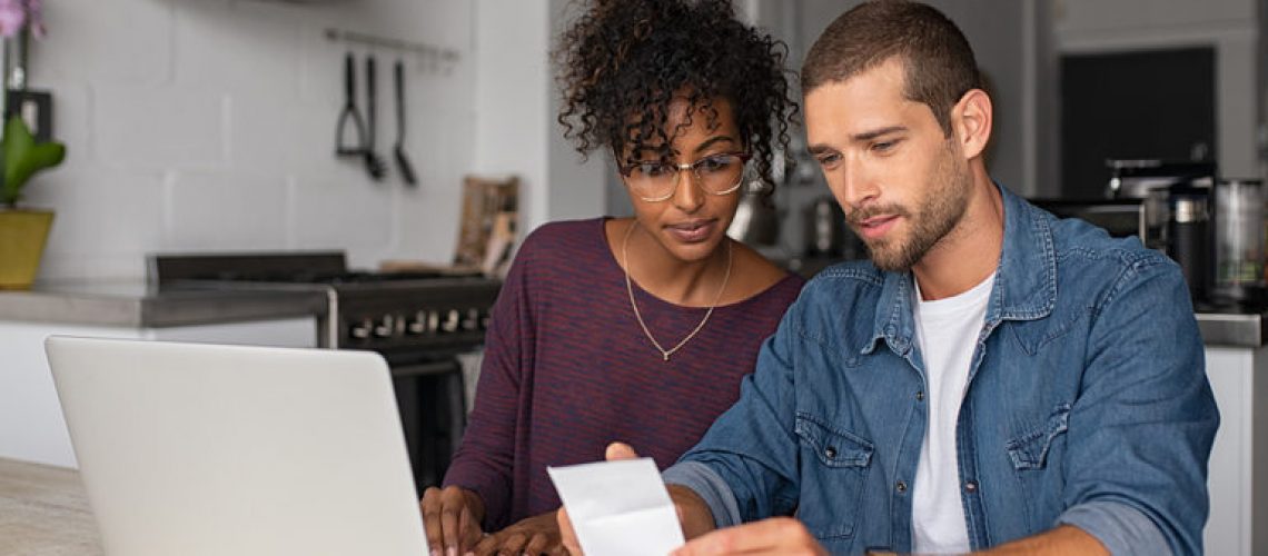 Young multiethnic couple checking bills while managing accounts on home banking app. Serious casual man and girl using laptop while looking at invoice and plan the budget to save.